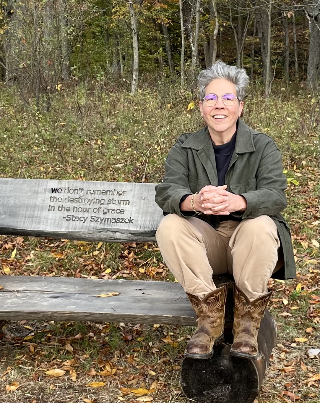A person, Stacy Szymaszek, with short gray hair and glasses sits smiling on a rustic wooden bench outdoors in autumn. They are wearing an olive green jacket, tan pants, and brown cowboy boots. They are smiling with their hands clasped loosely in front of them. Behind them is a wooded landscape with thin trees and fallen leaves in shades of brown, yellow, and orange. The backrest of the bench is engraved with text written by Szymaszek: “we don’t remember / the destroying storm / In the hour of grace."