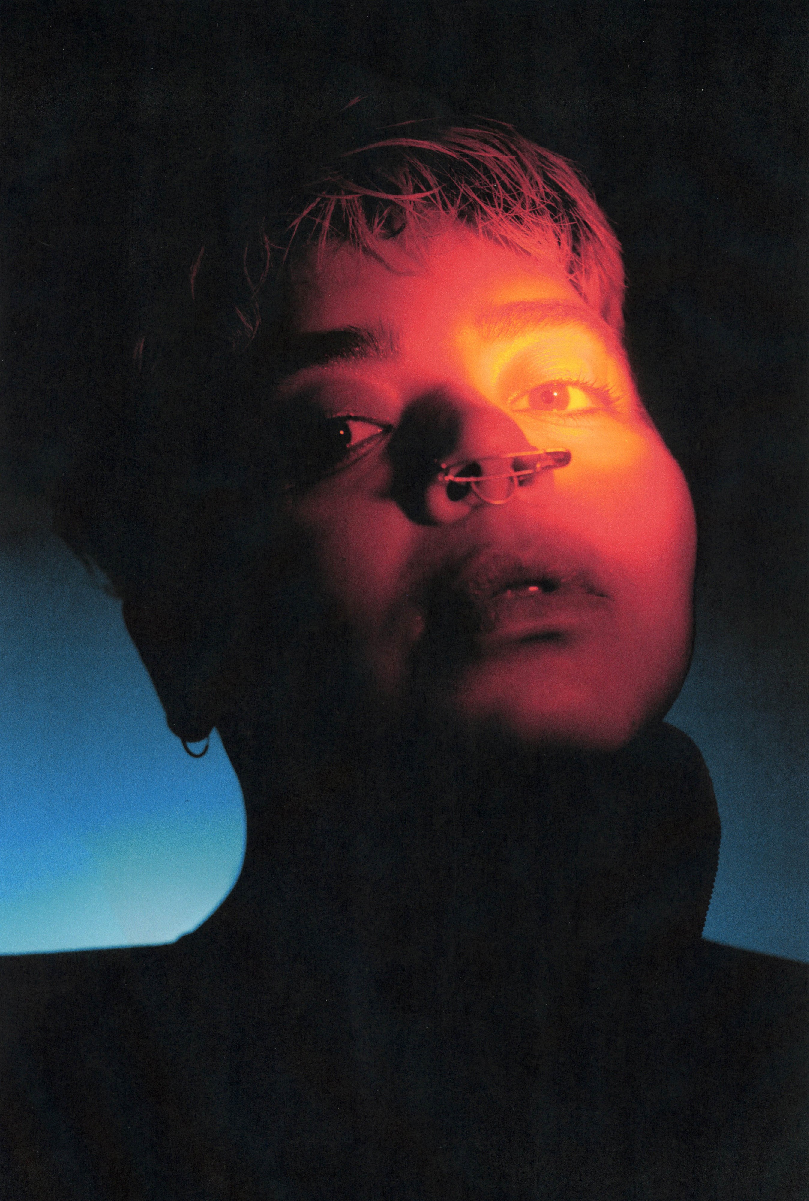 stefa marin alarcon gazes toward the camera with intent softness as a pool of warm orange and red light illuminates their left eye and dissipates across their face. Their nose pierced with a safety pin and a septum ring while the shadow of their ear discloses a small round earring. They are lit from behind with a small blue light deep in the image’s background, most of their head and collar left in dark silhouette.