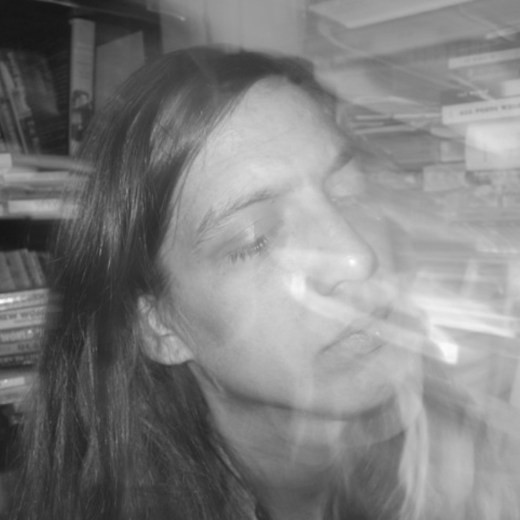 In this black and white portrait, Steph Gray looks away from the camera. Bookshelves are visible behind her, and her face is obscured by traces of light