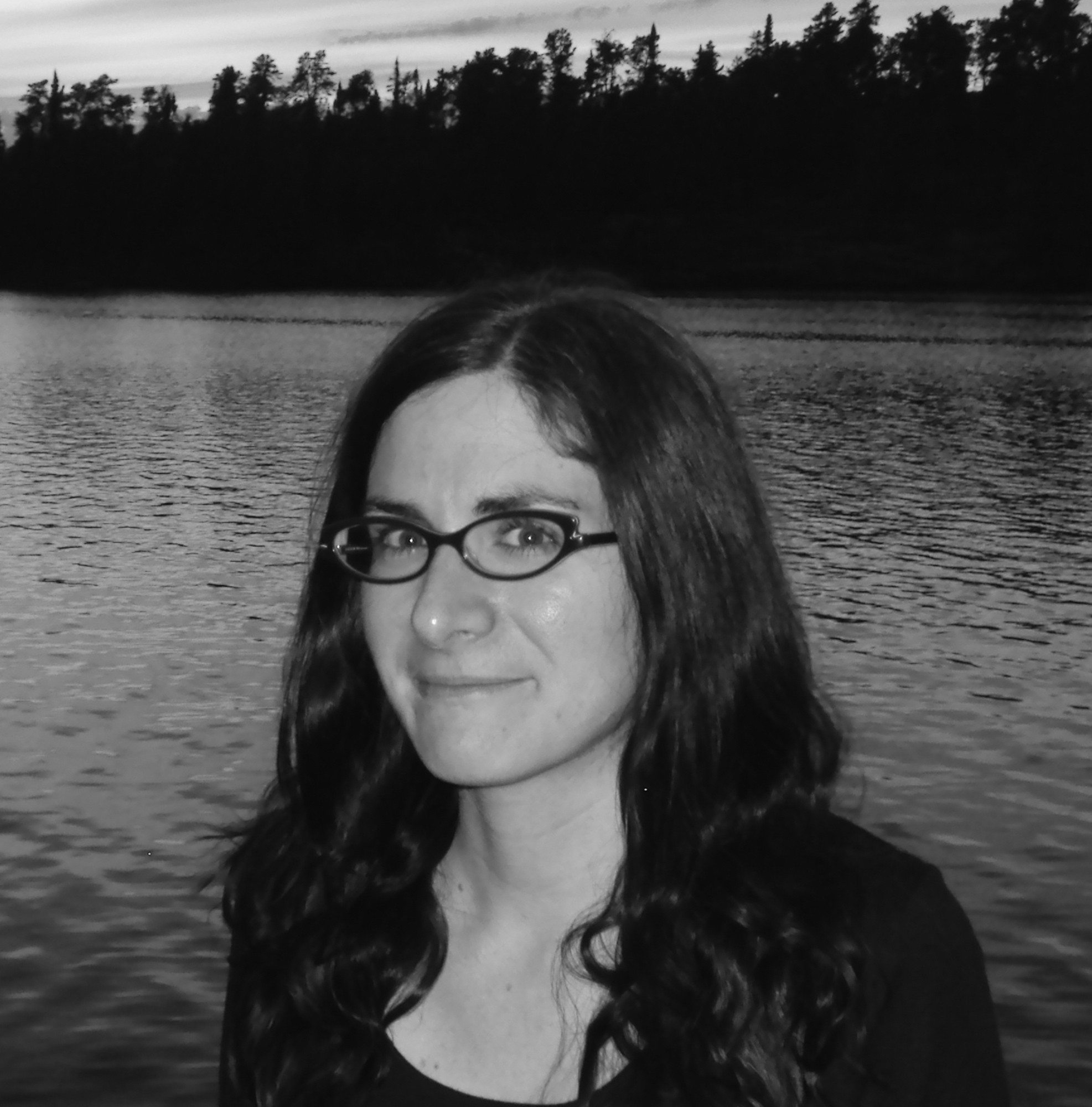 In this black and white portrait, Sylvia Matas is standing in front of a body of water, with a treeline visible on the opposite shore.