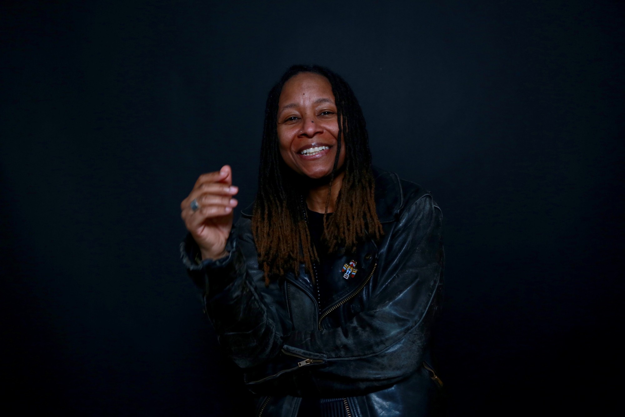 t’ai freedom ford is wearing a black leather jacket and standing in front of a black background. She is looking at the camera and smiling, her hand lifted in front of her and out of focus, as if in motion.