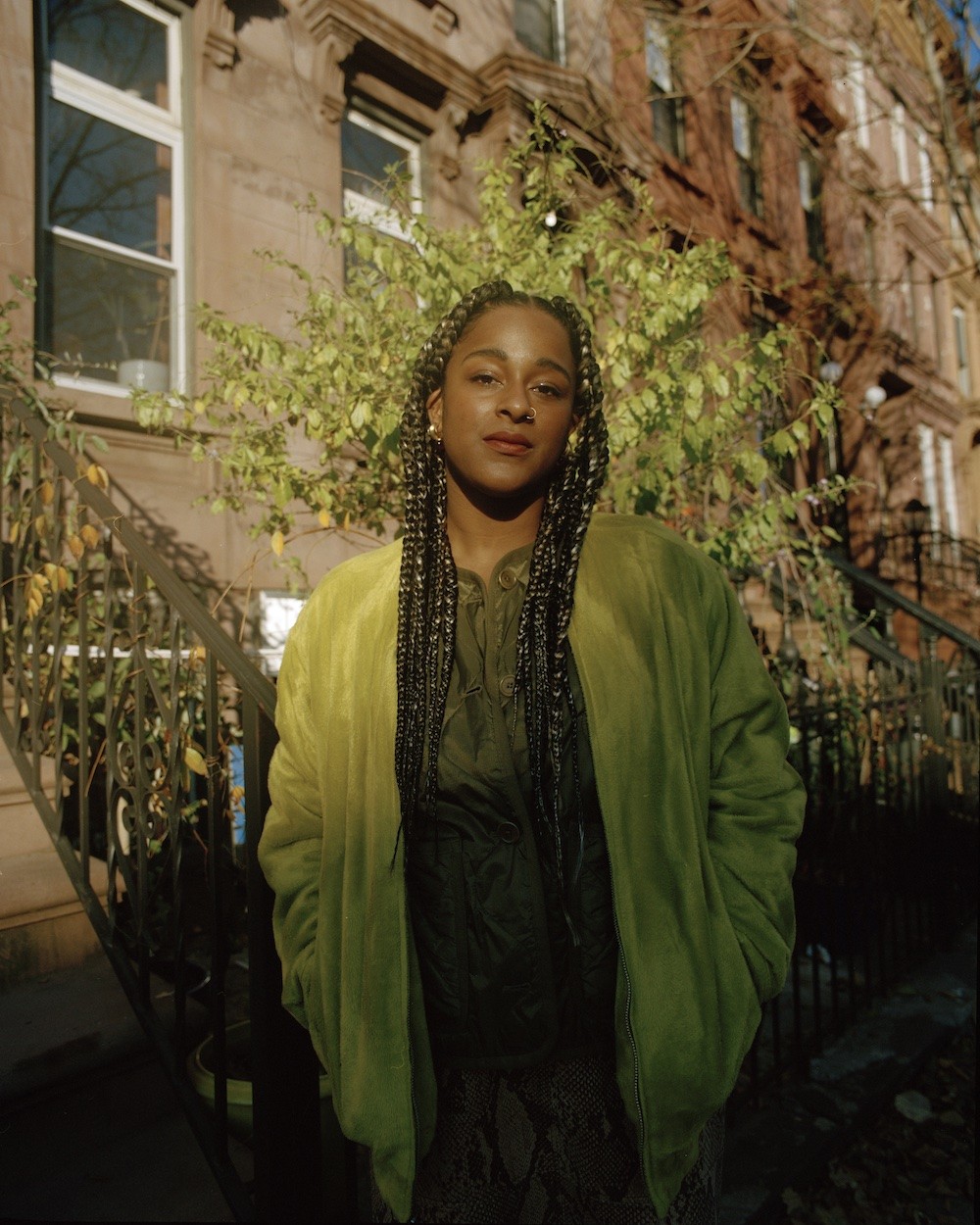 Taja Cheek is dressed in green, standing in front of a row of brownstones, caught between sunlight and shadows