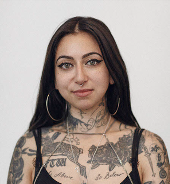 Tamara, a nonbinary person, with long brown hair, looks at the camera. They are standing in front of a white background, wearing a silver septum ring, silver necklaces, and a large silver hoop earrings. They have tattoos on their chest, neck, and upper arms and wear a black tank top.