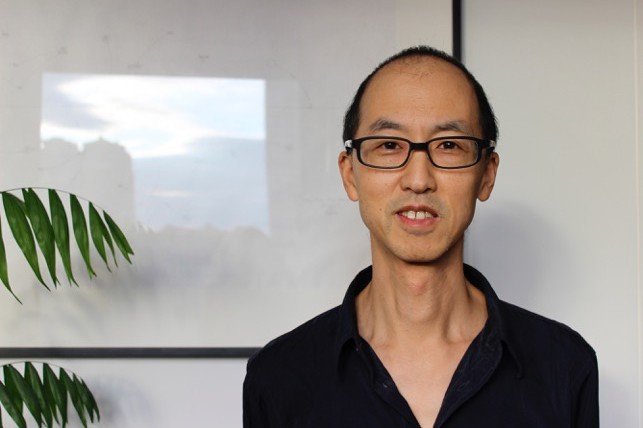 Tan Lin wears a black button up shit and black enamel glasses. He's standing in front of a blank whiteboard, a plant is visible at the edge of the frame