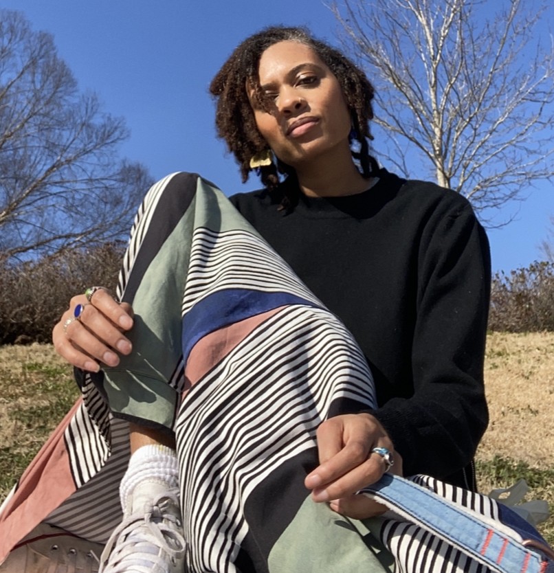 Tash Nikol sits outside on the ground wearing a black turtleneck, white converse, and a long skirt, striped with multiple colors. The sky is blue, the grass is yellow, and the trees have no leaves.
