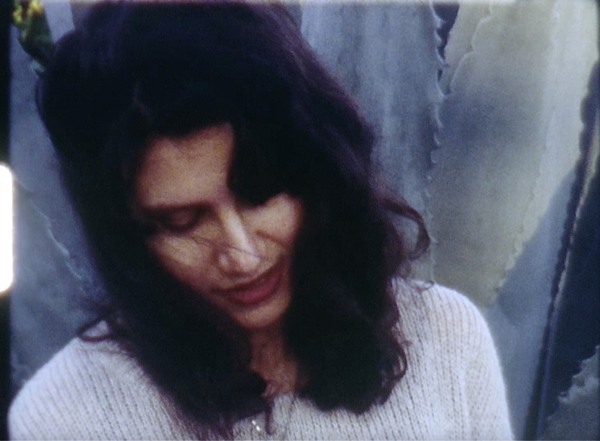 Tatiana, a femme presenting non-Black Latinx person with shoulder length dark brown hair and dark red lipstick, is outside on a windy, late afternoon, looking downwards and smiling slightly. They are wearing a knitted oatmeal colored sweater and standing in front of a massive agave plant. The image is a film still from a Super 8mm film: it is grainy, and a sprocket is visible on the left hand side.