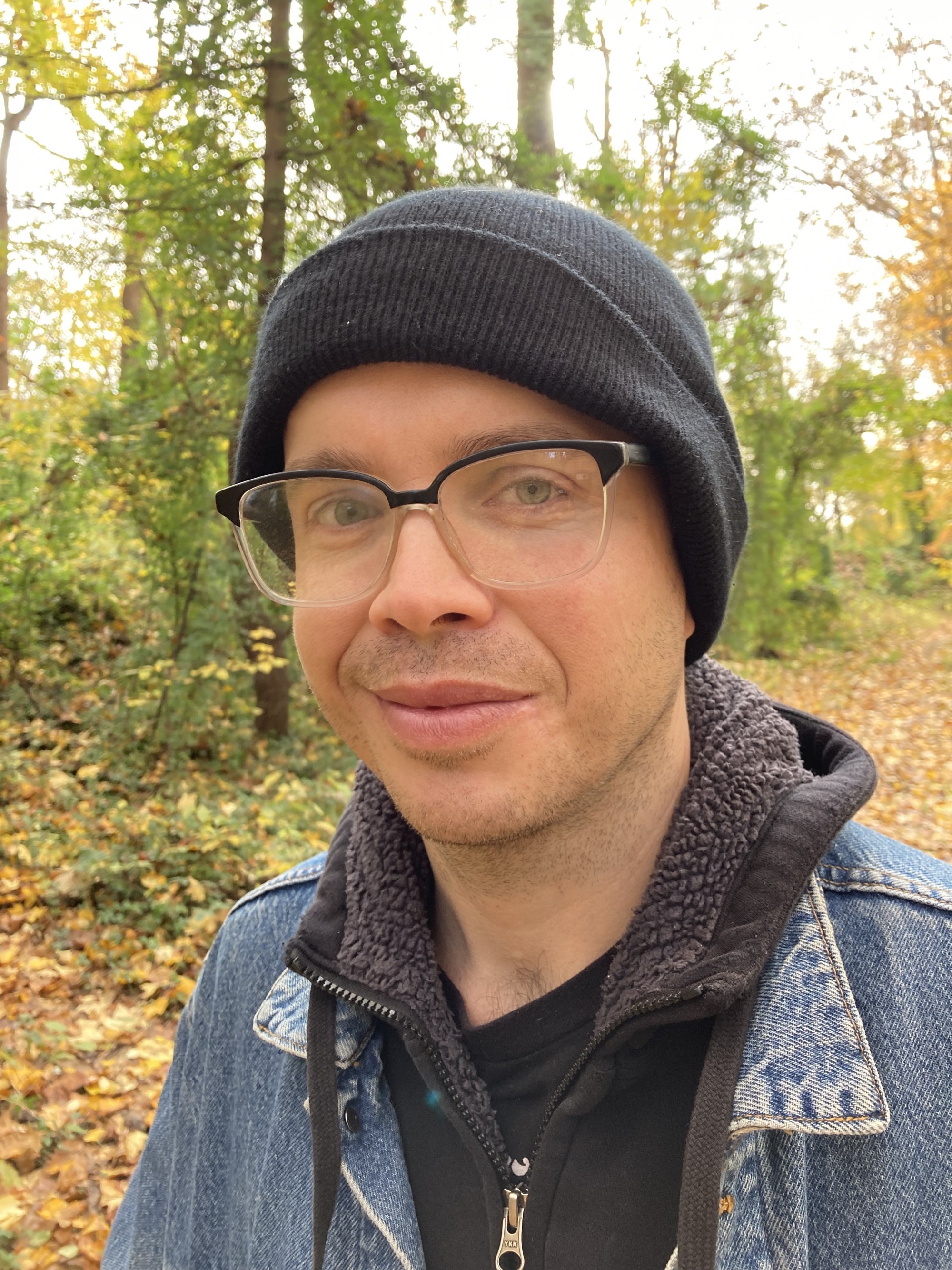 Ted Rees wears glasses and a beanie, standing in the woods. Leaves are on the ground and he's smiling.