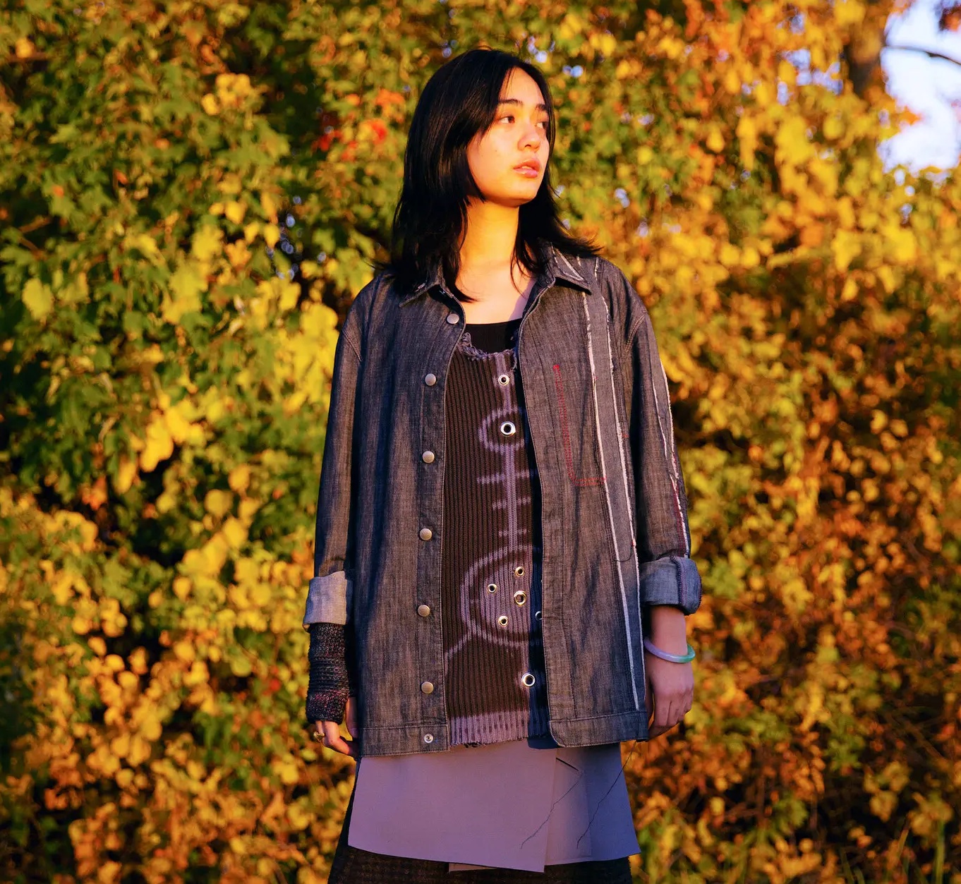Teline is standing outside in the sun, out of focus green leaves visible behind them, looking into the distance