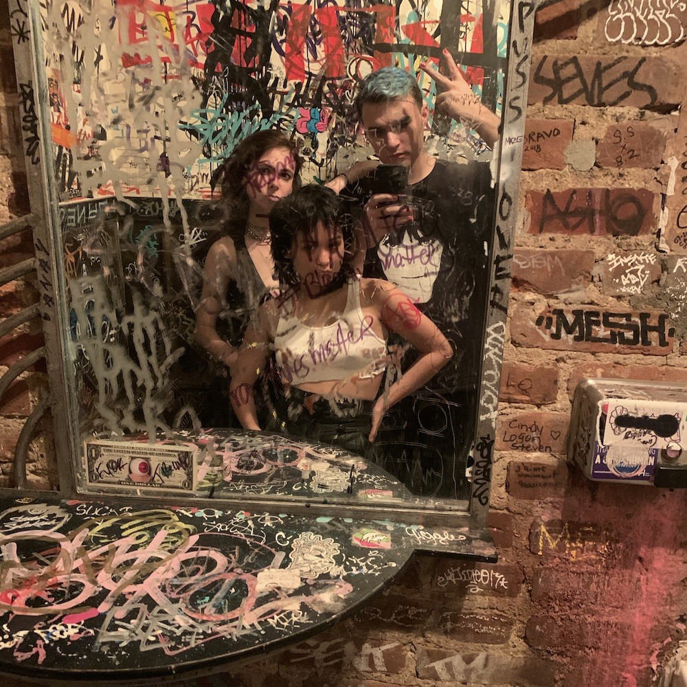 In this mirror selfie, members of The Anchoress Syndicate stand together in front of a heavily graffitied mirror on a heavily graffitied wall, each looking very cool.