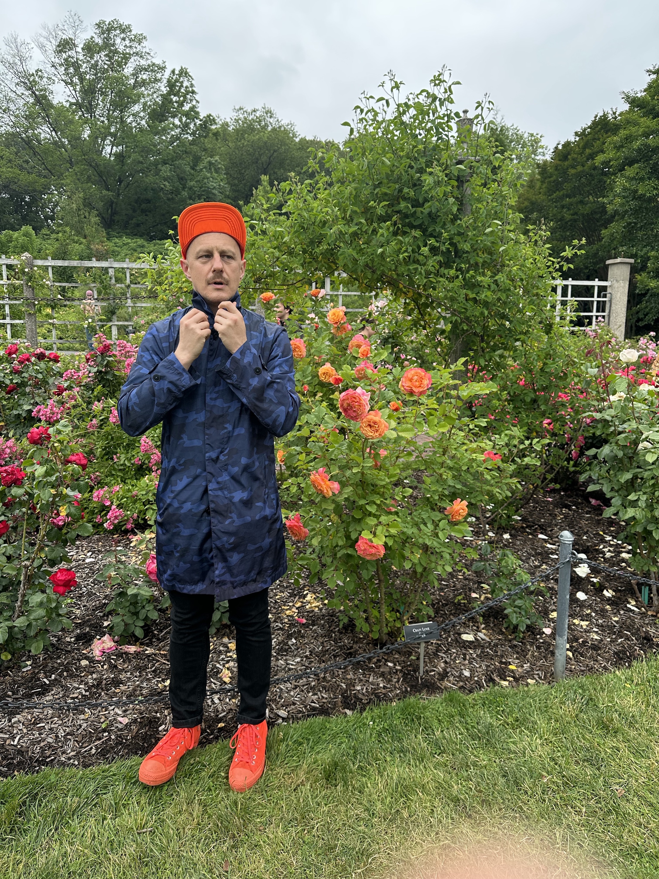 A white middle aged man with a mustache stands in a garden. He is wearing a bright orange cap on his head that almost matches the color of his shoes. He is also wearing dark blue pants and a blue camouflage rain coat.  The expression on his face suggests he is kinda goofy.