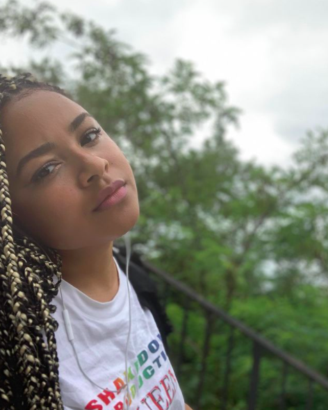 Tiana Reid stands outside by a railing, her head tilted slightly back, her hair in long braids, looking into the camera