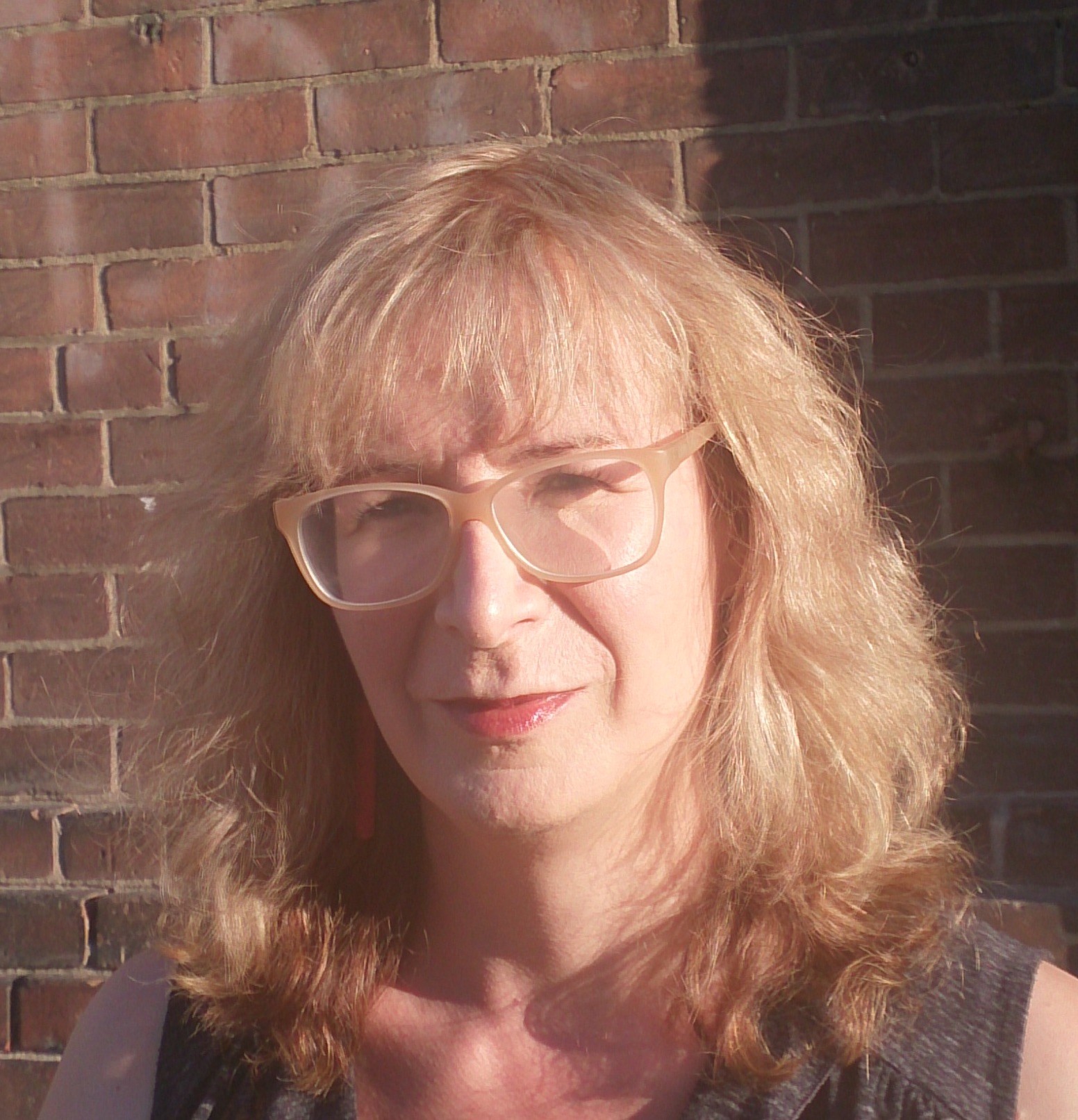 Trish Sala stands in the sun in front of a brick wall, wearing red lipstick and glasses