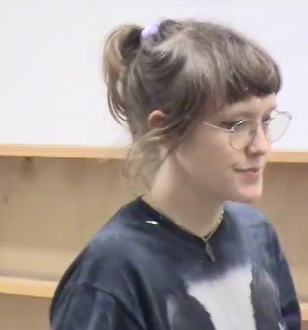 Tyler  a white queer with a loose ponytail and circular gold-rimmed glasses, walks in front of a bare bookshelf and a white wall in a video still. Her face is in profile. She wears a black and white shirt, a necklace with one half of a heart dangling from it, and a lavender hair tie.