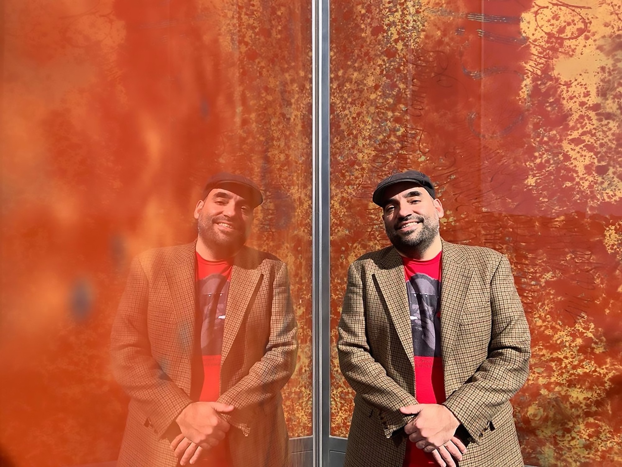 Urayoán stands in front of a red wall, wearing a hat, and a blazer over a red t-shirt. He is looking at the camera and smiling.