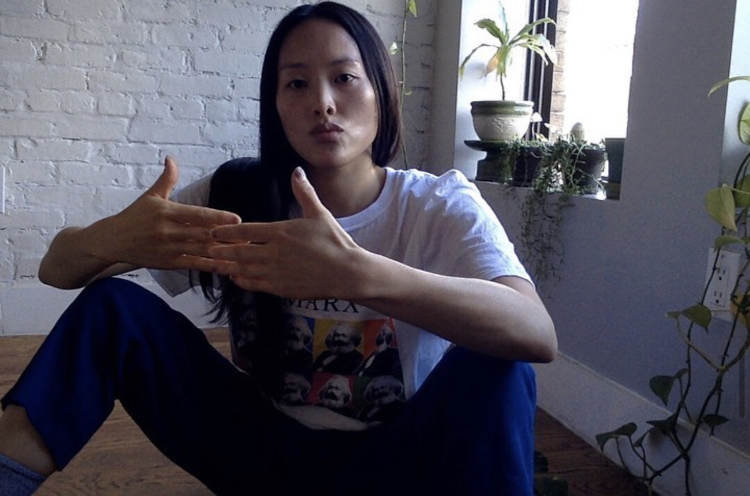 An Asian woman with long hair, arms raised making a gesture with her hands of convergence, and a white t-shirt with replicated images of Karl Marx sits on a wooden floor in front of a white brick wall and some plants on a windowsill.