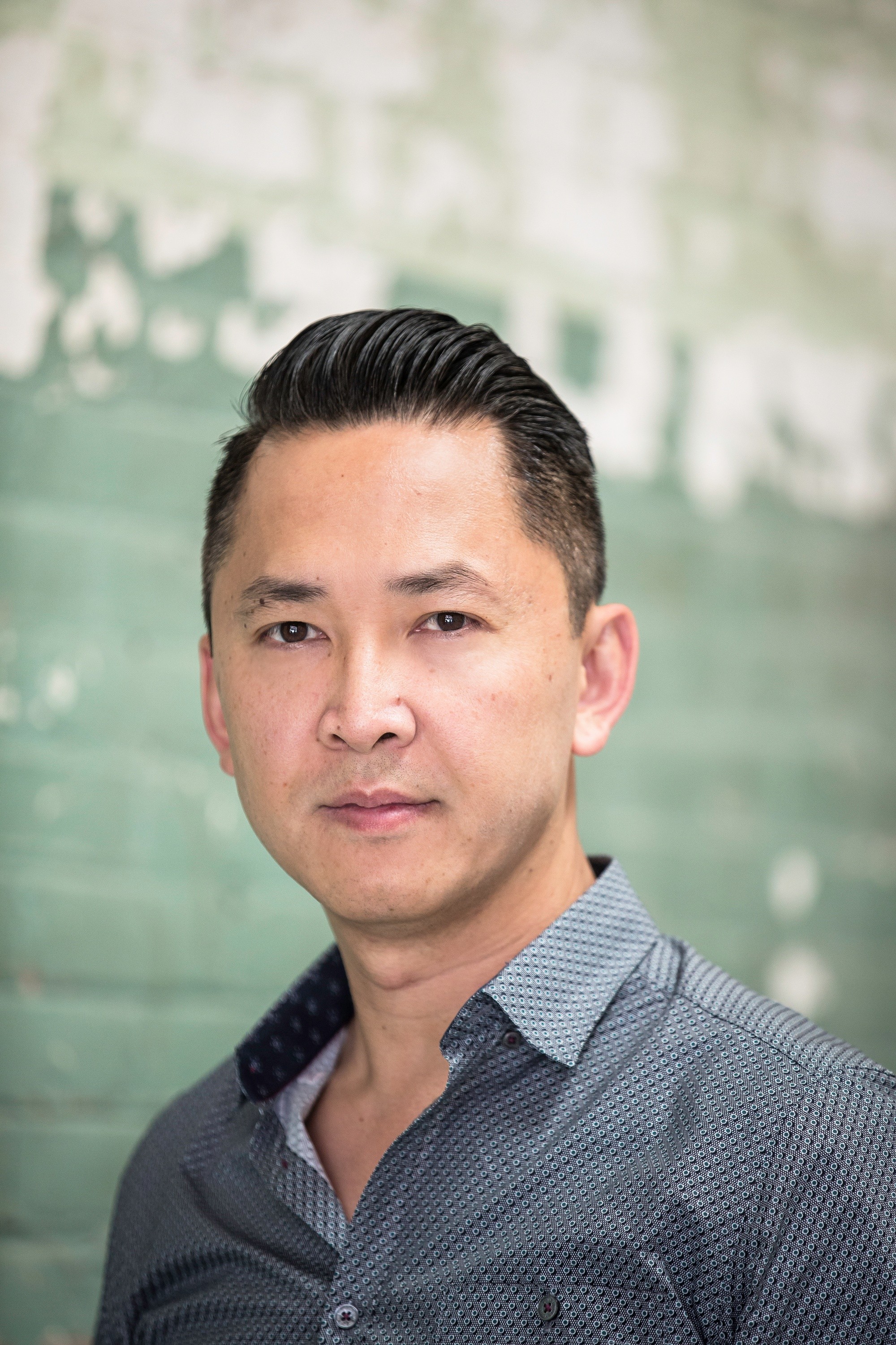 Viet Thanh Nguyen stands in front of a green brick wall, wearing a button-up shirt, looking at the camera.