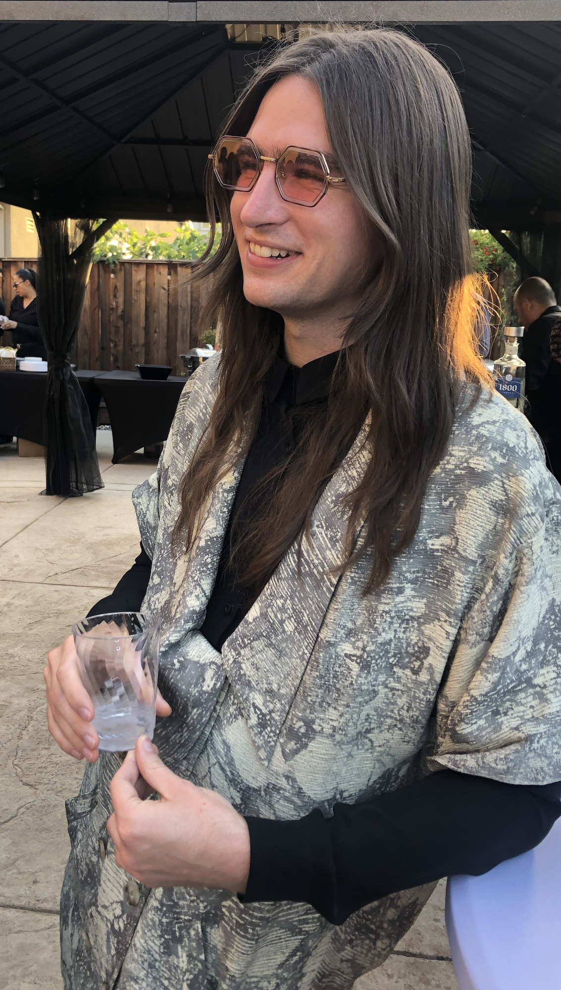 A woman wearing sunglasses and an oversized blazer is smiling and holding a glass of water.