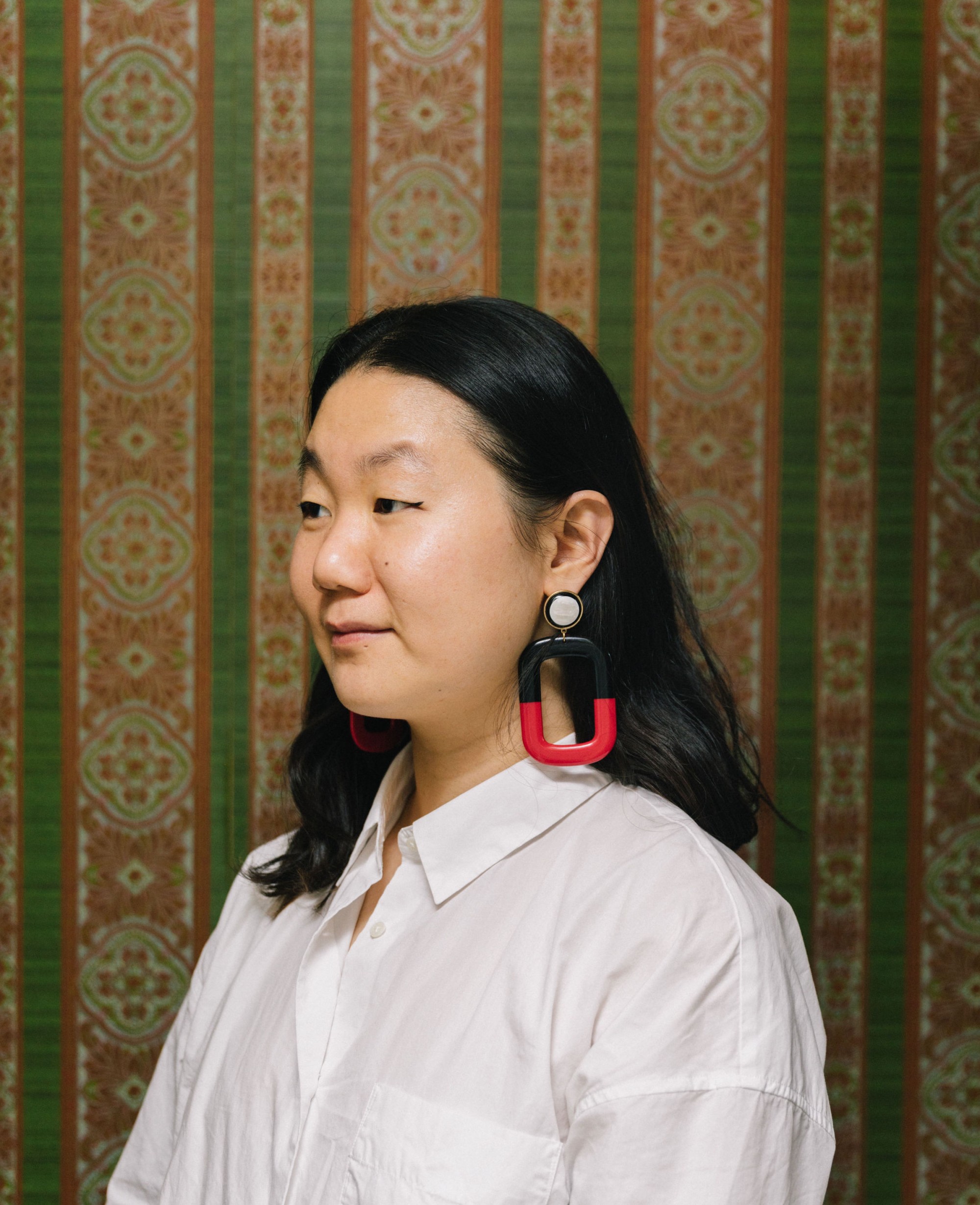 Wendy Xu stands in front of green and orange vertical striped wallpaper, wearing a white shirt and large red and black earrings, looking to the side.
