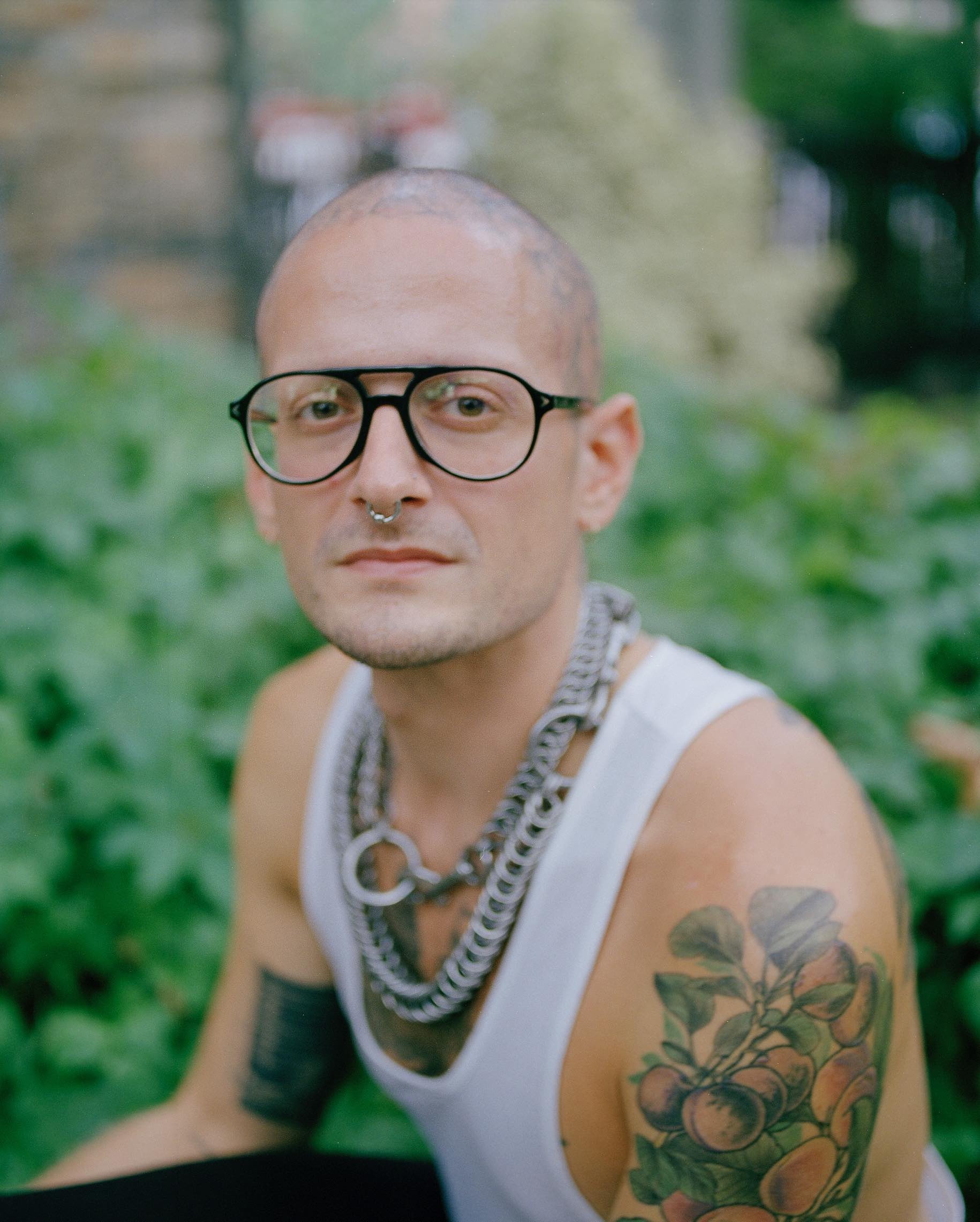 Will, a white nonbinary person with a shaved head, is shown in a portrait-oriented large-format film photograph cropped roughly from the chest up. They are facing the camera at a three-quarter angle. Will is wearing a loose white tank top, two heavy chain necklaces, heavy black enamel glasses in an aviator style. Their septum is pierced and they have a tattoo of a peony that covers their whole head. The depth of field in the photograph is focused tightly on Will so the background is blurry, but greenery is visible.