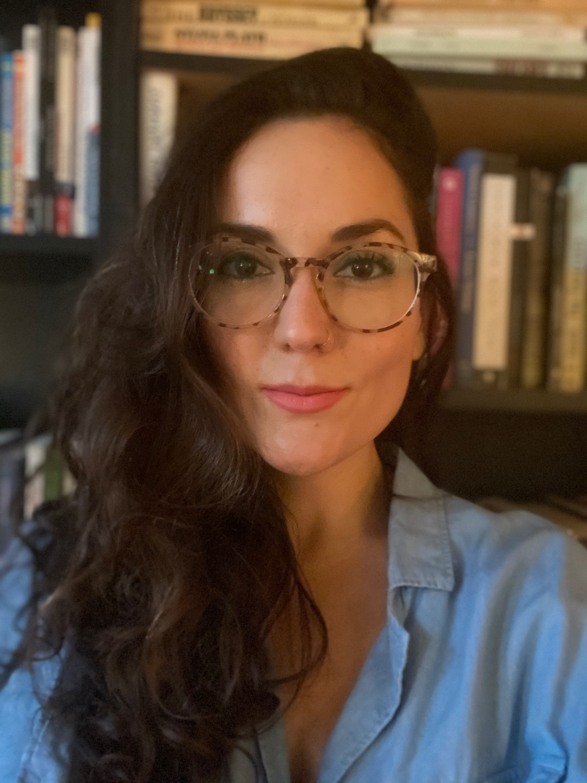 In this photo, Ximena Keogh Serrano faces the camera, wearing glasses and pink lipstick, in front of a bookshelf