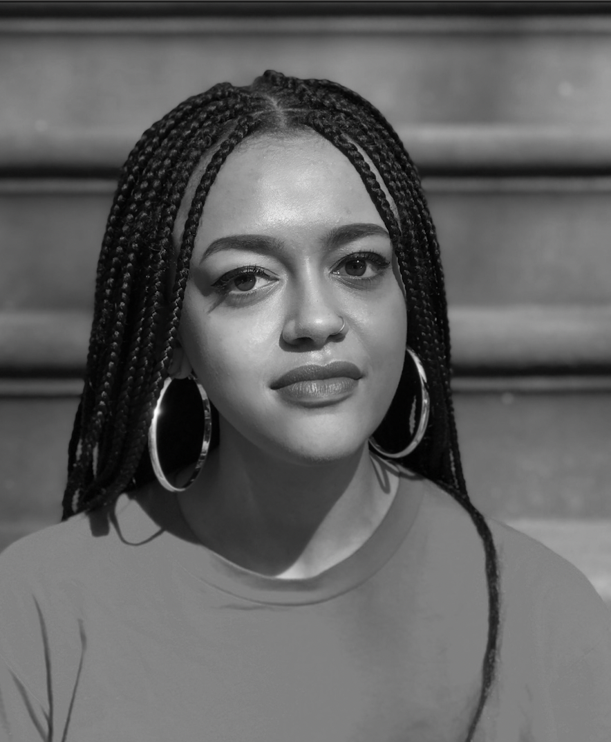 Yasmina in a black and white close-up portrait photograph. She is a black woman with box braids and silver hoop earrings, wearing a t-shirt with stairs in the background.