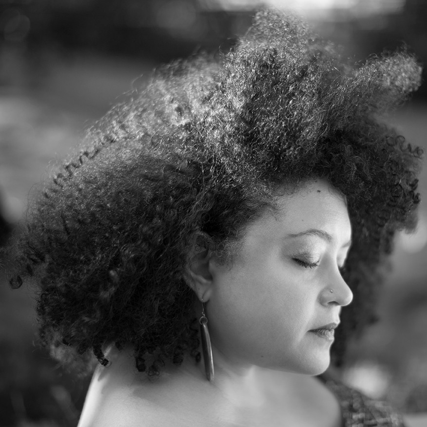 Yesenia Montilla is featured in a square-cropped black-and-white portrait in profile that is softly lit by natural lighting. The photo is cropped from the shoulders up. She is wearing long pendant earrings and her hair frames her face.