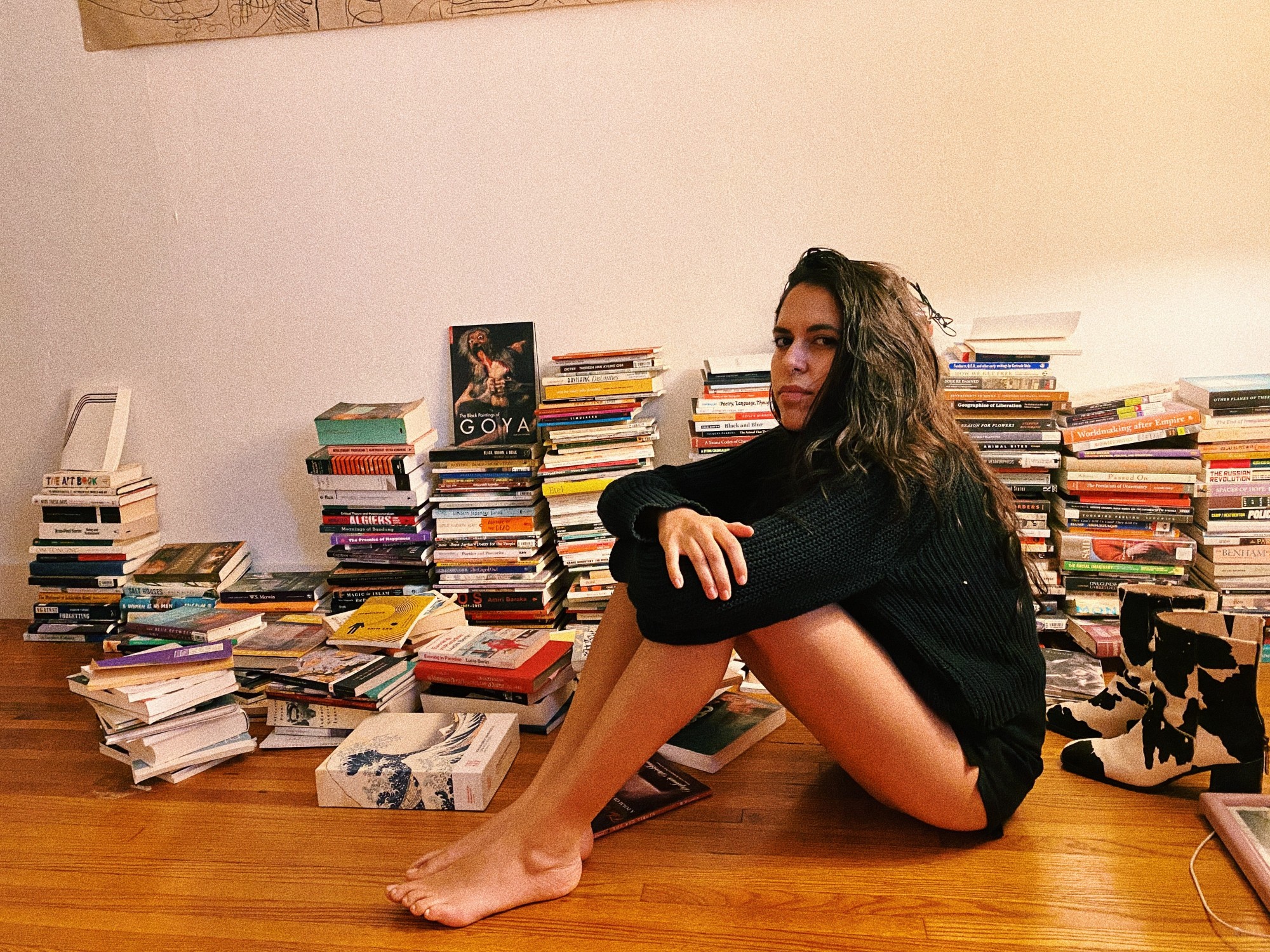 Zaina Alsous sits on the floor in a black sweater in front of a big pile of books