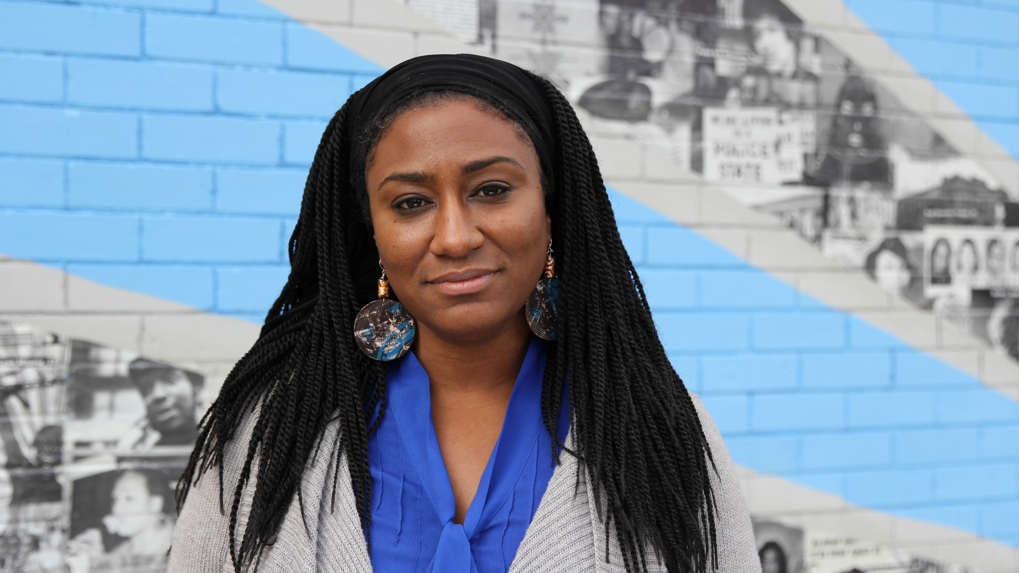 Zenzele Isoke looks directly into the camera, smiling, standing in front of a muraled wall, just out of focus.