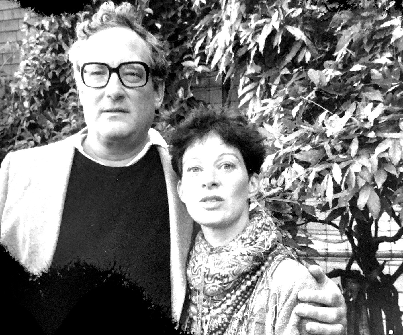 A close-up, black-and-white photograph of Kenward Elmslie holding Ann Lauterbach to his side. They're standing at the side of a house, in front of some trees, both looking a bit bewildered at the camera.
